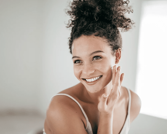 A Smiling Woman In A White Tank Top With Her Hair Tied Up And A Makeup-free Face Gently Brushes The Side Of Her Face With One Hand.