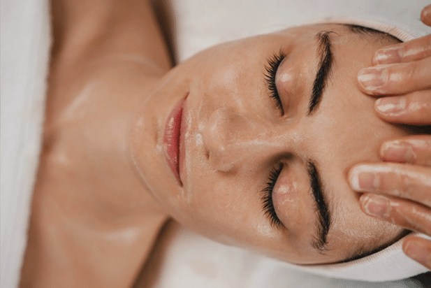 Close Up Of A Woman‘s Face And Shoulders While She‘s Lying Down With Her Eyes Closed And Someone‘s Fingertips Apply A Facial Serum To Her Face.