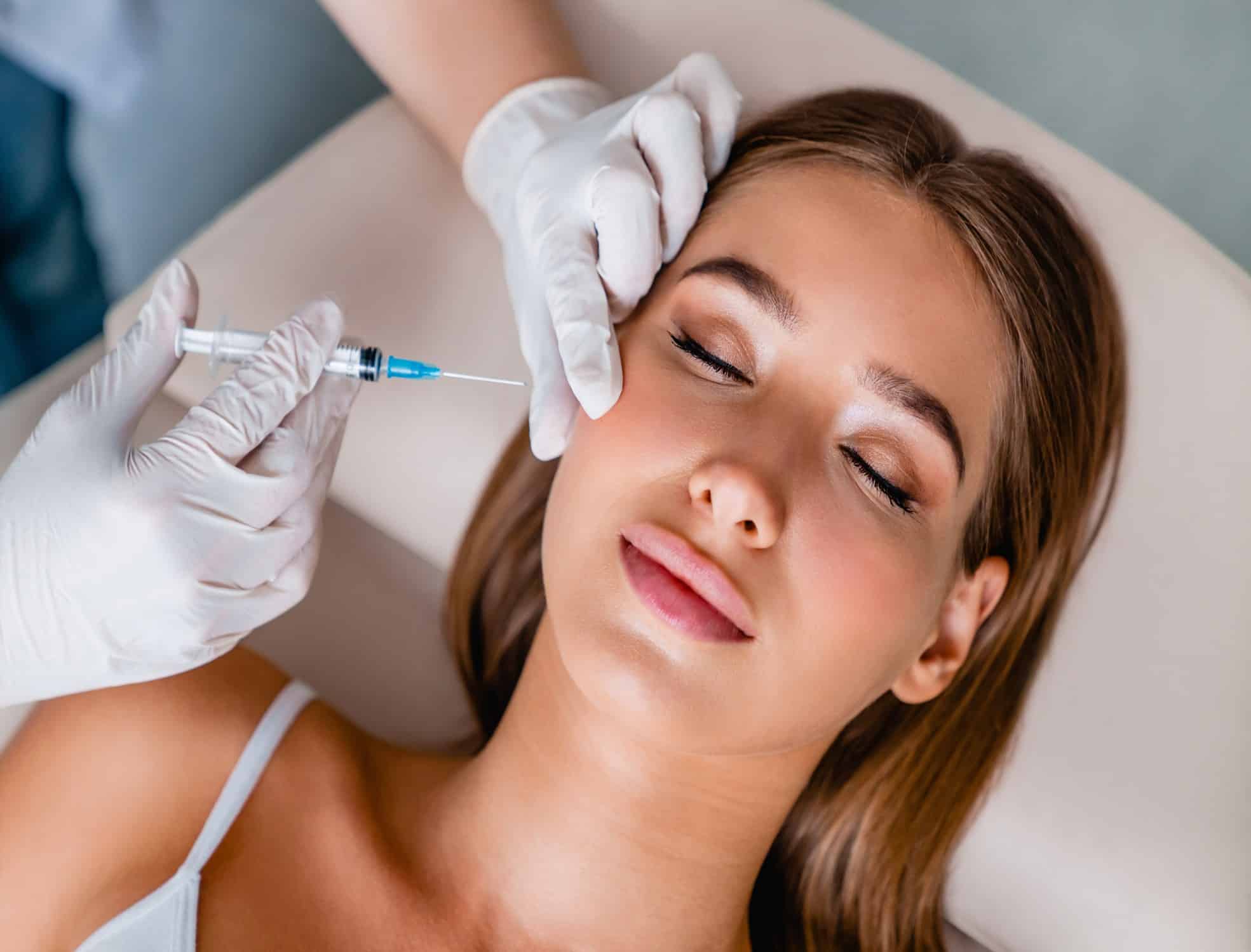 A Woman Lying Down With Her Eyes Closed While An Injection Specialist Places One Gloved Hand On The Side Of Her Face And Uses The Other To Inject A Treatment Into Her Cheek.