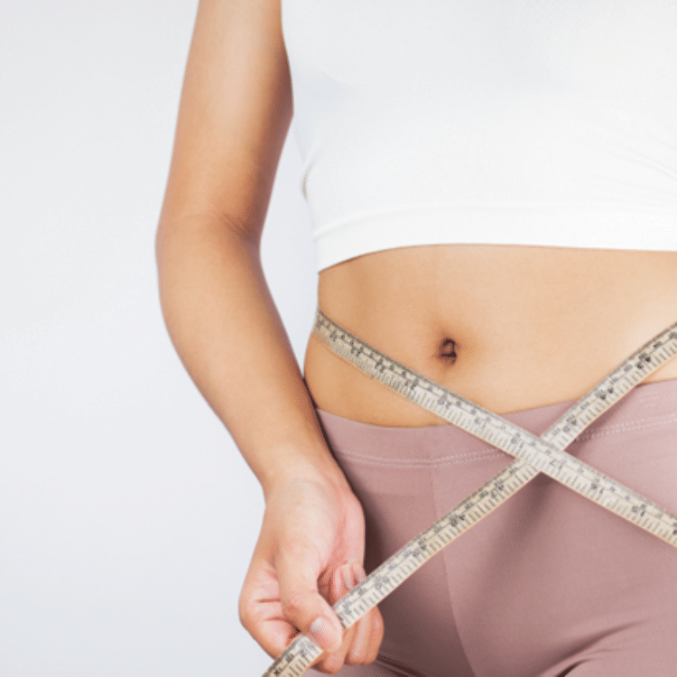 A woman‘s torso and right arm in a white tank top and mauve bottoms as she crisscrosses a measuring tape across her stomach.
