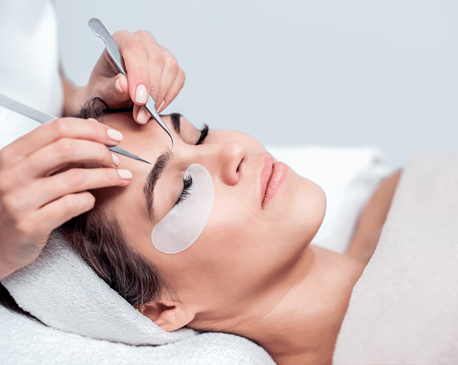 A woman lies on her back with her eyes closed and a headband around her head with an eye pad under her right eye while a woman uses tools to apply lash extensions to her right eyelid.