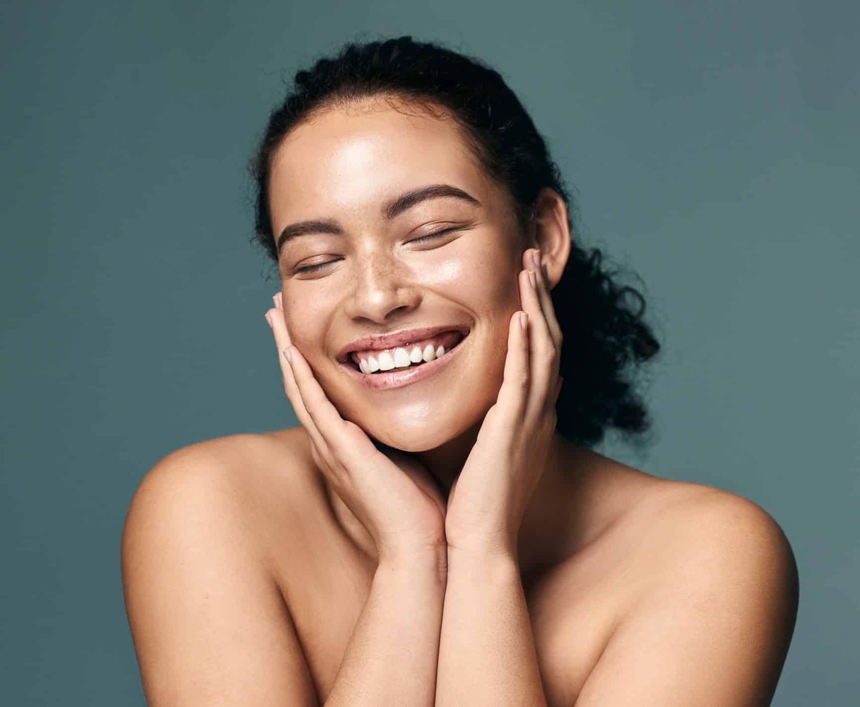 A fresh-faced smiling woman with bare shoulders, freckles across her nose, and dark curly hair cups the sides of her face with both hands.
