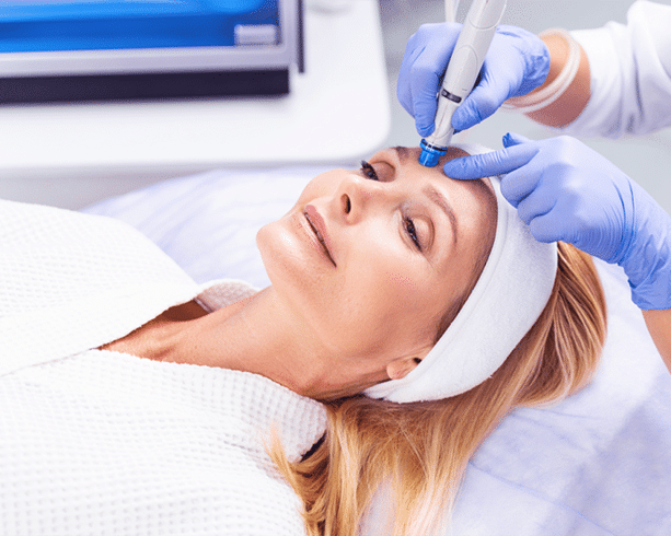 A mature woman in a white robe lies on her back with a headband around her head while two gloved hands use a microdermabrasion tool on her forehead.