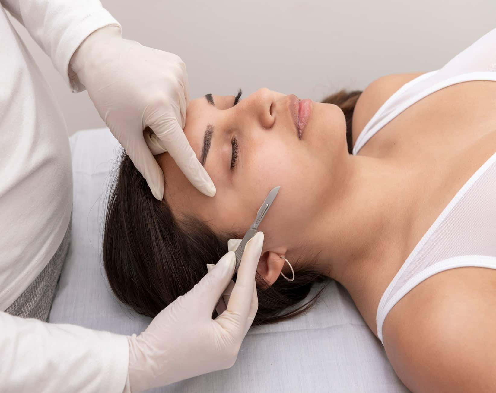 A woman lying on her back with her eyes closed while one gloved hand from another person touches the side of her face and another uses a dermaplaning blade on her cheek.