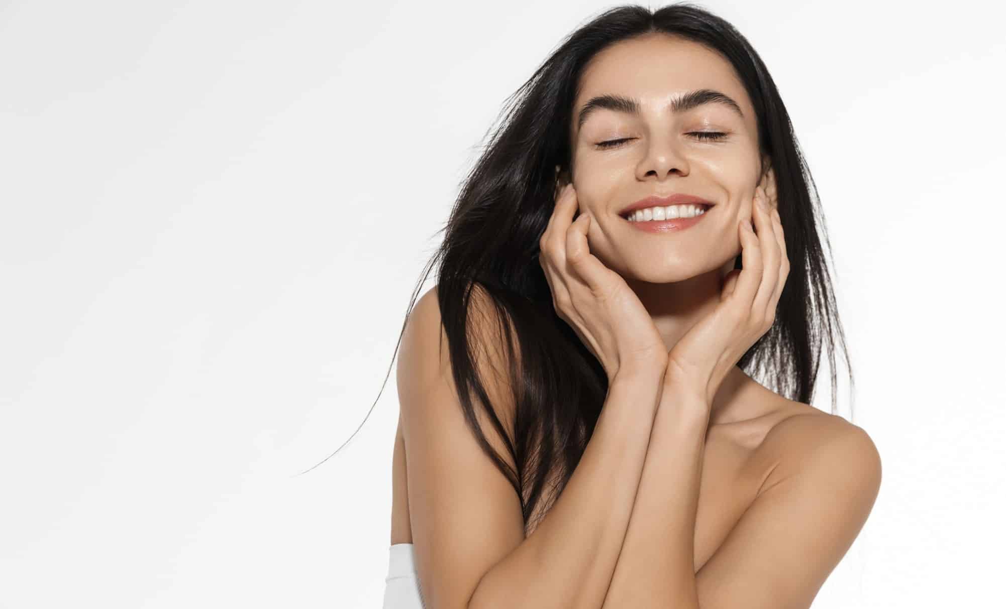 A smiling brunette woman with bare shoulders and dewy skin smiles with her eyes closed and cups both sides of her face with her hands.