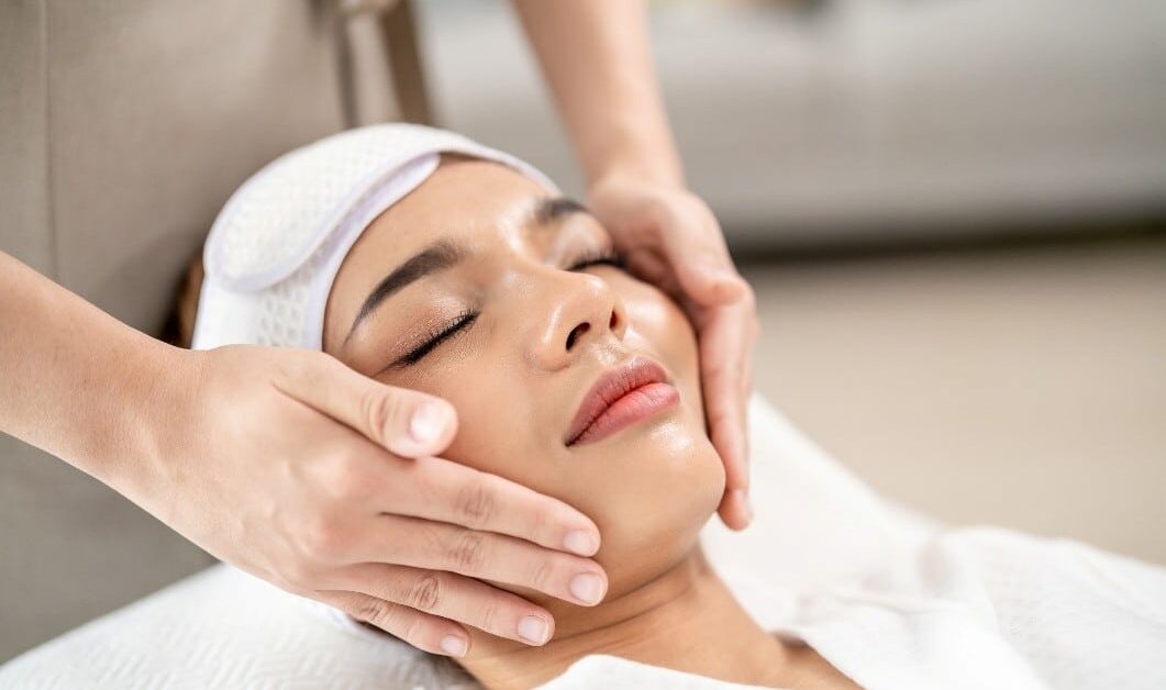 A woman with eyes closed and a white headband receives a relaxing facial massage from a spa professional.