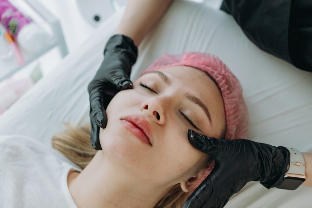 Overhead view of a woman wearing a pink hairnet receiving a professional facial treatment from a practitioner wearing black gloves.