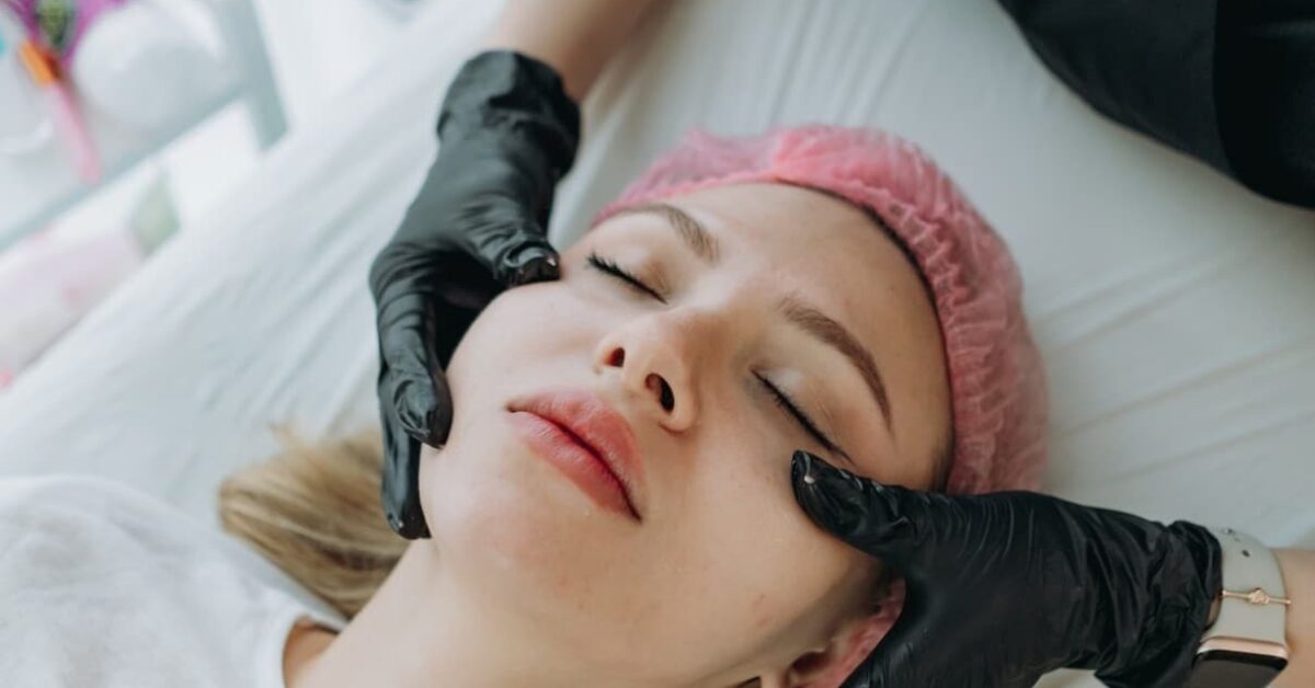 Overhead view of a woman wearing a pink hairnet receiving a professional facial treatment from a practitioner wearing black gloves.